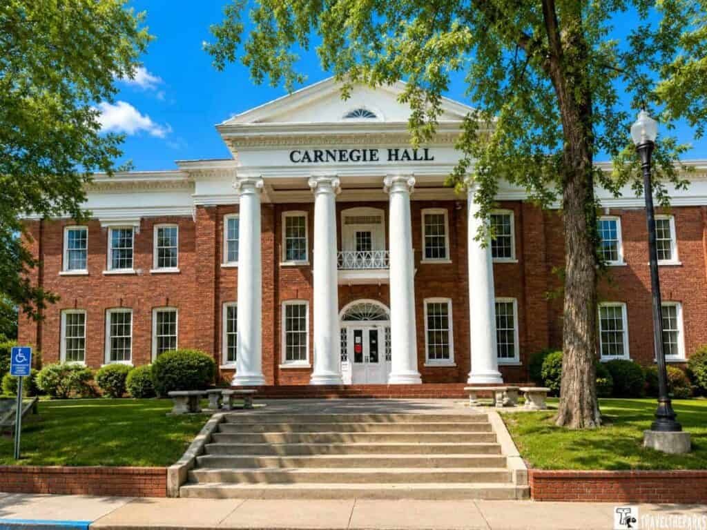 A classic brick building named Carnegie Hall, featuring white columns and large windows, surrounded by green lawns and trees under a blue sky.