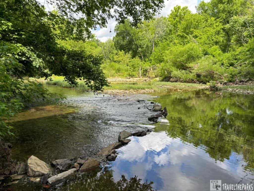 A rocky path leads into a serene river surrounded by lush green trees and underbrush at Stones River National Battlefield.

