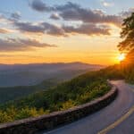 A winding road curves around a wooded hillside at sunset, with rolling mountains and a colorful sky in the background.