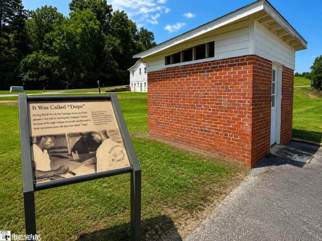 A historical marker sign in front of a small brick building at the Tuskegee Airmen National Historic Site, depicting two individuals looking at documents.

