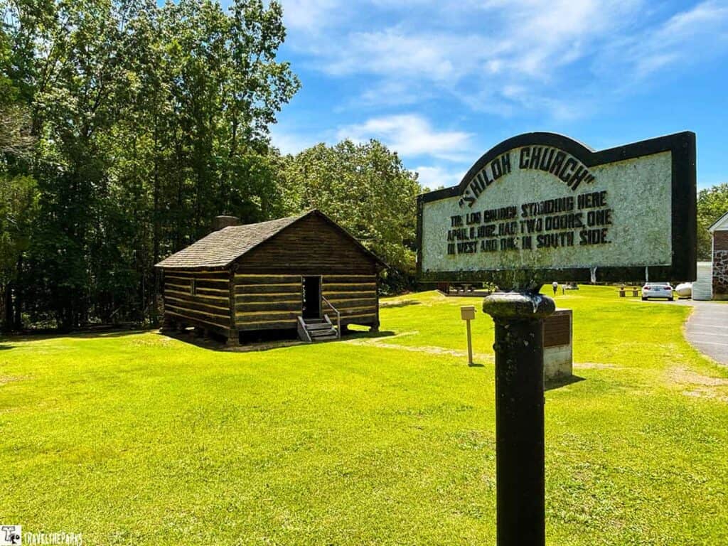 A log church named Shiloh Church stands on a green lawn, accompanied by a black sign detailing its history. Trees and a clear blue sky are in the background.