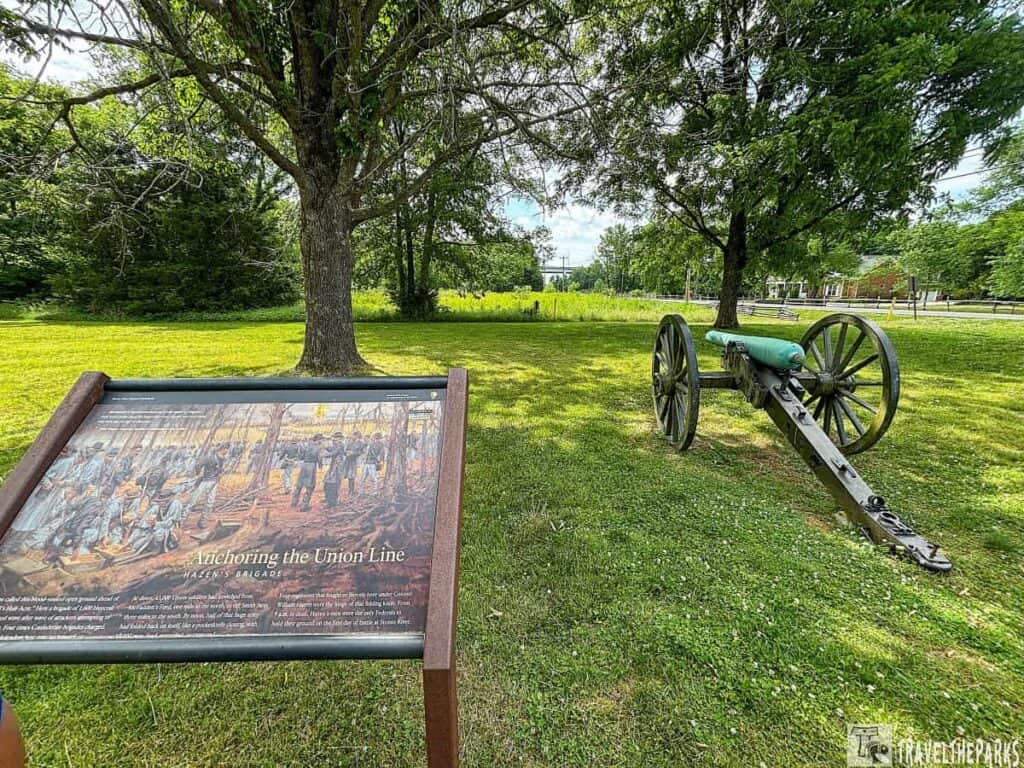 A vintage cannon on grassy ground next to an informational sign about Union soldiers, surrounded by greenery and trees at Stone River National Battlefield.