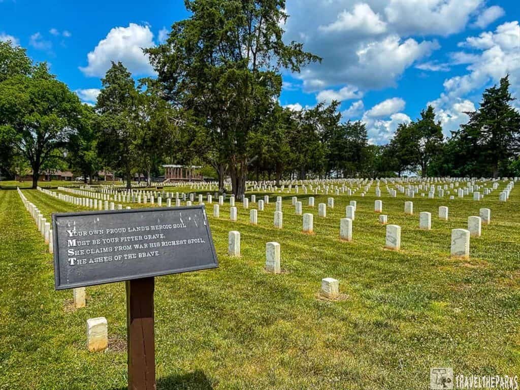 A cemetery with rows of white gravestones set in green grass, a black plaque in the foreground with engraved text, and trees and blue sky in the background.

