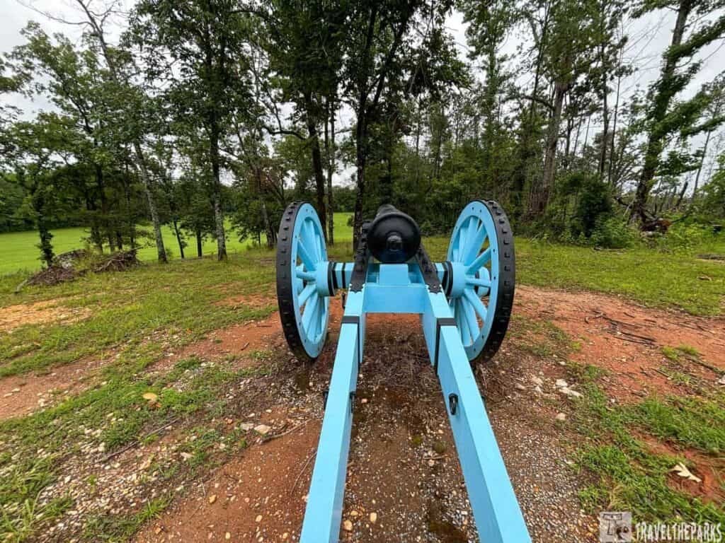 A blue cannon with black wheels sits on a dirt path surrounded by greenery, with trees in the background at Horseshoe Bend National Military Park.