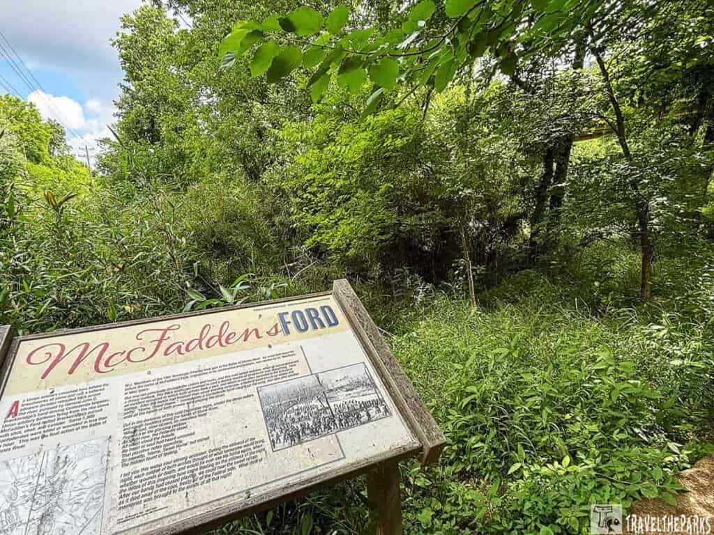 A wooden sign reading "McFadden's Ford" stands amidst dense greenery and trees at the Stones River National Battlefield, under a partly cloudy sky.

