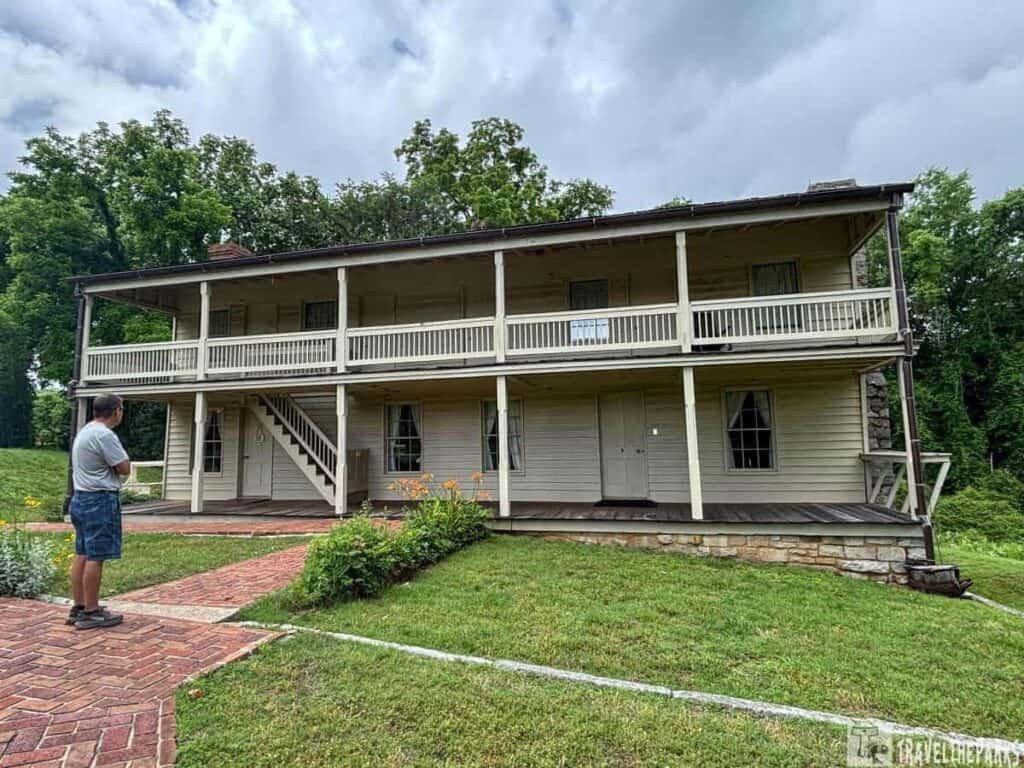 Two-story Surrender House at Fort Donelson with a balcony, surrounded by a green lawn and flower beds, partially cloudy sky, and a person standing in front.