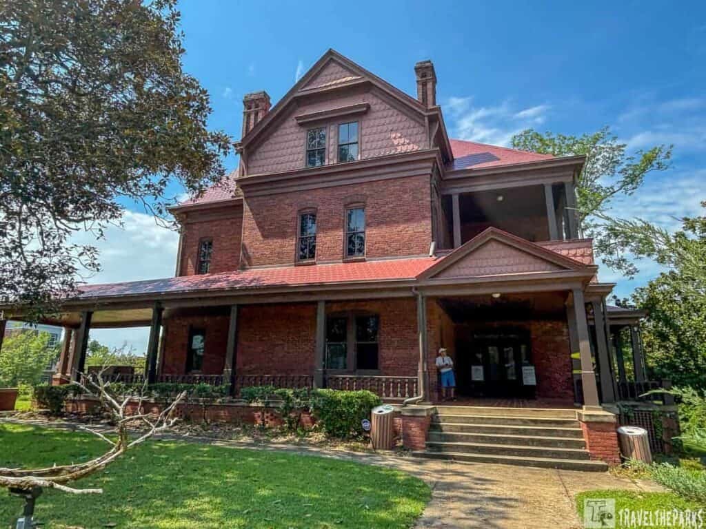A historic Victorian house known as "The Oaks," Booker T. Washington’s home at Tuskegee Institute, with a red brick exterior, gabled roof, and lush surrounding greenery.