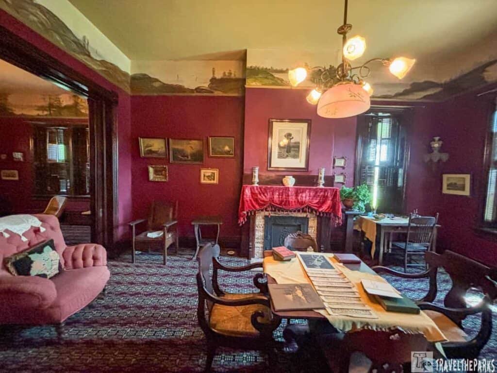 Interior of the parlor at Booker T. Washington’s home, featuring pink furniture, maroon walls with framed art, a fireplace draped in red, and a table covered with papers.
