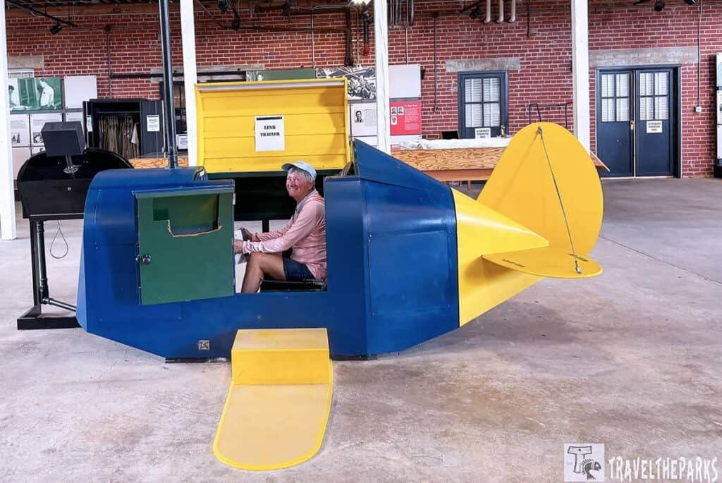 A woman smiles while seated in a blue and yellow miniature airplane cockpit inside a historic aviation exhibit, with a green control panel and a yellow storage box labeled "LINK TRAINER" in the background.