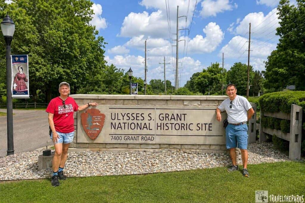 Two people pose beside a sign for the Ulysses S. Grant National Historic Site, surrounded by greenery and a blue sky with clouds.