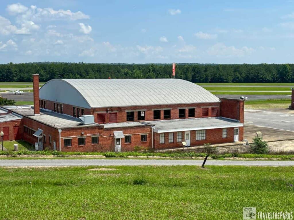 Red brick building with an arched gray roof at the Tuskegee Airmen National Historic Site, surrounded by green grass and an airstrip in the background.