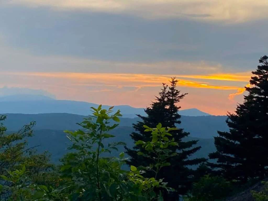 A sunset landscape with distant mountains, evergreen trees in the foreground, and a colorful sky featuring orange and yellow hues.