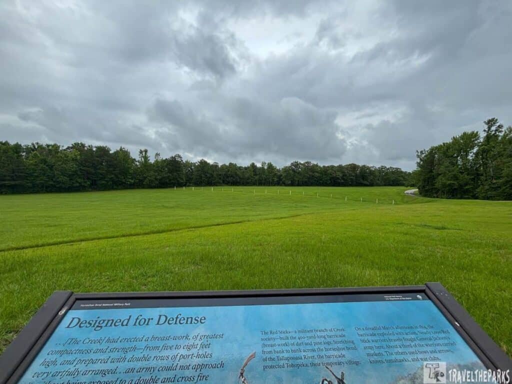 Landscape view at Horseshoe Bend National Military Park showing a green field with white posts marking the Red Stick barricade and a sign providing historical context, under an overcast sky.