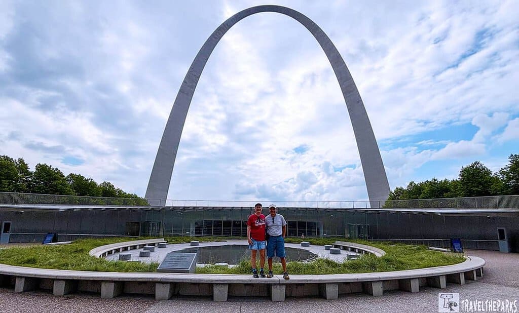 Two men stand in front of the Gateway Arch in St. Louis, surrounded by greenery and a circular water feature under a cloudy sky.