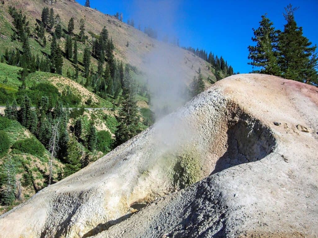 A volcanic mound at Lassen Volcanic National Park with rising steam, surrounded by green hillsides and clear blue sky.

