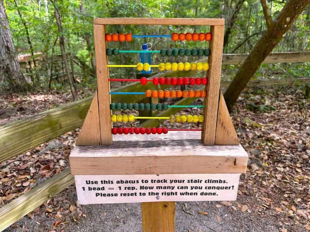 a colorful wooden abacus positioned on a wooden stand in a natural outdoor setting at Devil's Millhopper Geological State Park. The abacus consists of several rows of colorful beads in varying shades, including red, orange, yellow, green, and blue, arranged on horizontal bars.