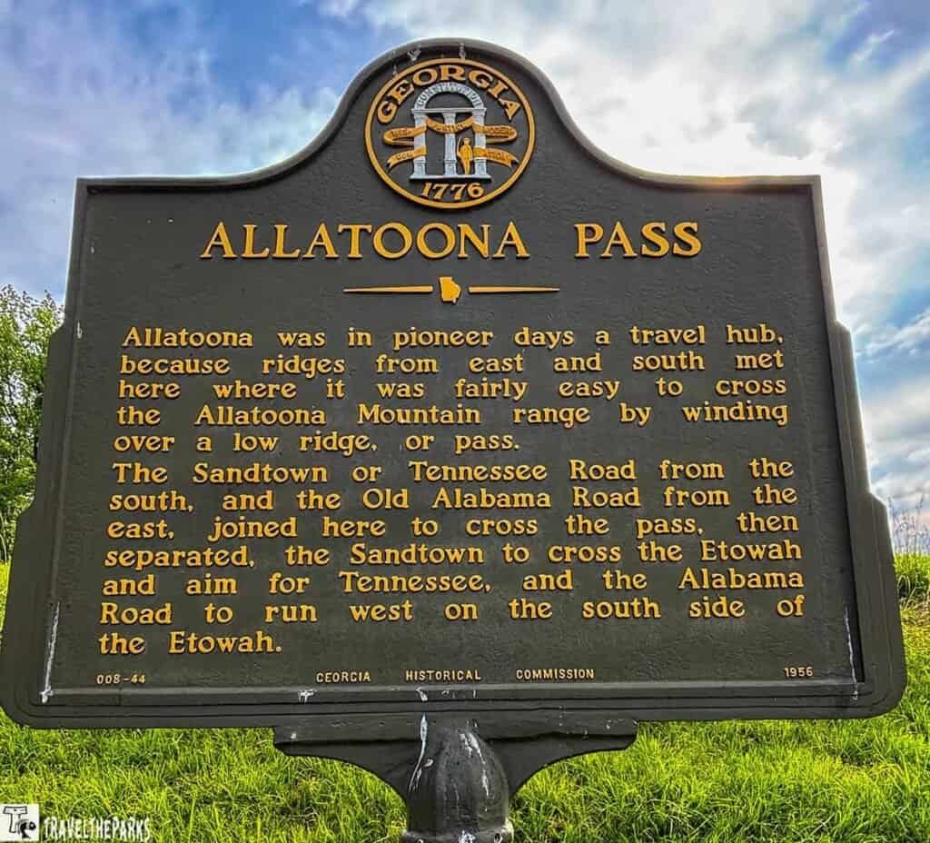 A black historical marker titled "Allatoona Pass" in Georgia, featuring gold text and the state seal at the top, provides a historical account of the location’s significance as a travel hub. Transcribed Text: ALLATOONA PASS Allatoona was in pioneer days a travel hub, because ridges from east and south met here where it was fairly easy to cross the Allatoona Mountain range by winding over a low ridge, or pass. The Sandtown or Tennessee Road from the south, and the Old Alabama Road from the east, joined here to cross the pass, then separated, the Sandtown to cross the Etowah and aim for Tennessee, and the Alabama Road to run west on the south side of the Etowah. 008-44 GEORGIA HISTORICAL COMMISSION 1956