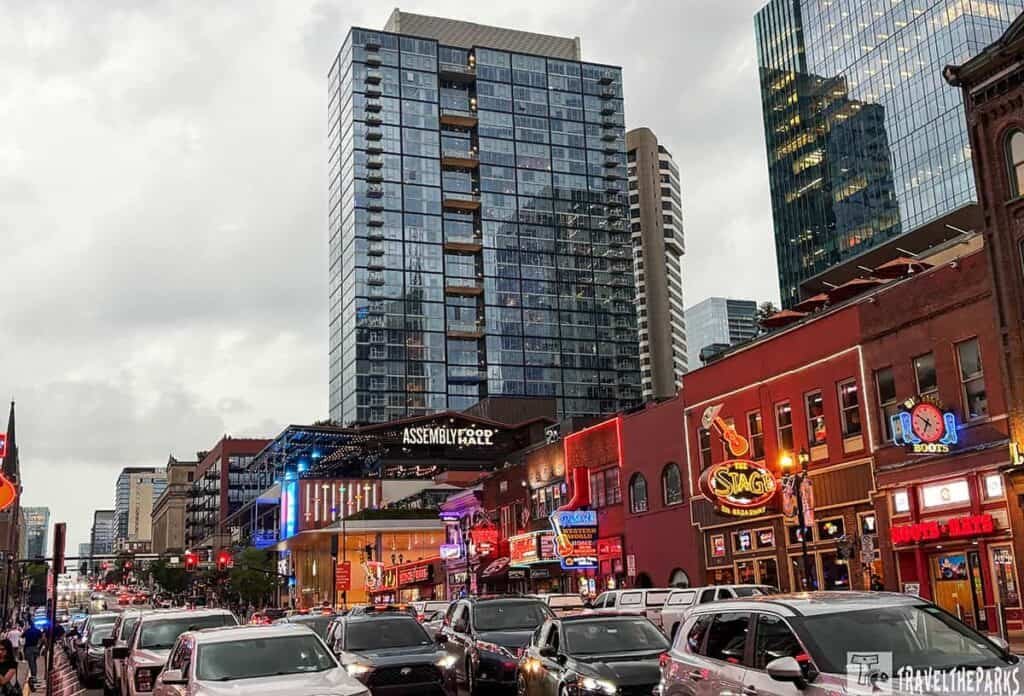 Urban Nashville Honly Tonk street scene at dusk with a tall glass skyscraper, busy traffic, and illuminated neon signs from nearby establishments.

