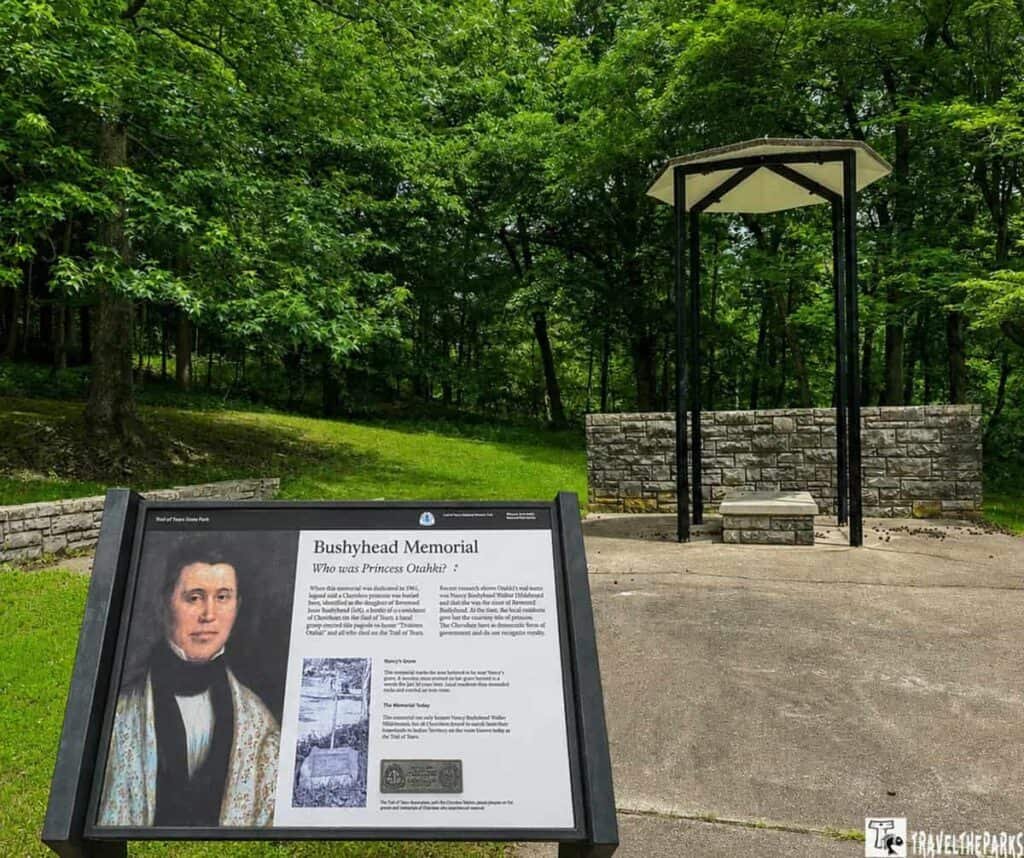 The image shows the Bushyhead Memorial at Trail of Tears State Park, featuring an information sign with a historical portrait and descriptive text. A stone platform and wall are visible behind the sign, with trees providing a lush background.