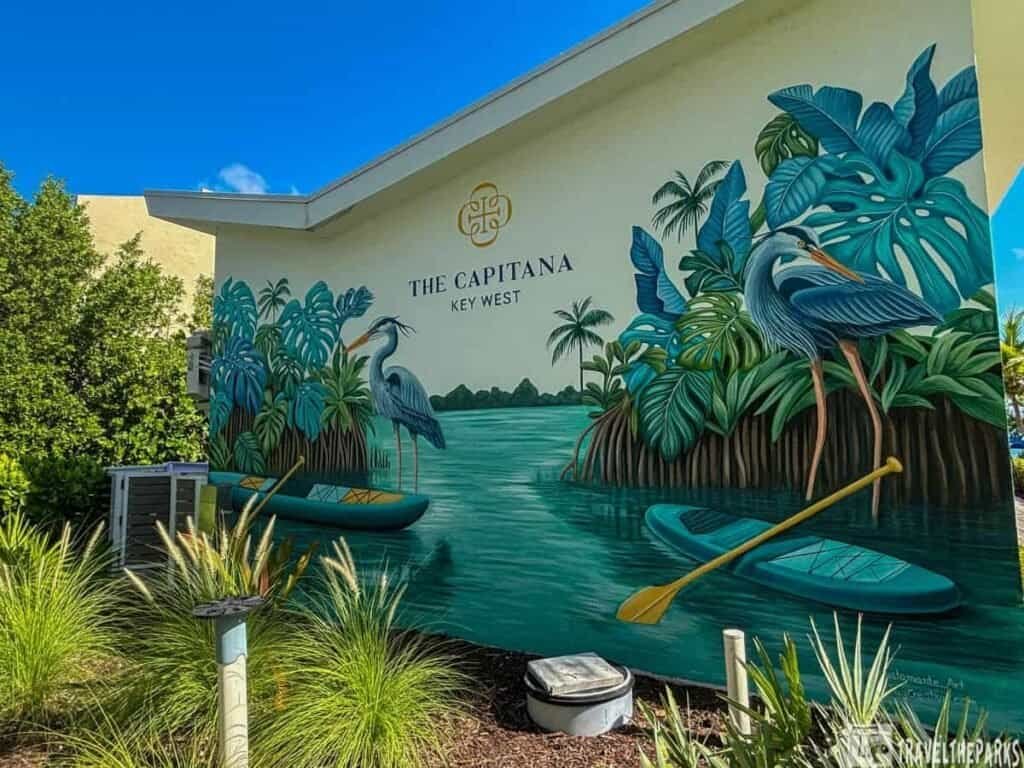 A mural on a building wall featuring "The Capitana Key West," depicting two herons, tropical foliage, and colorful kayaks on calm water with a blue sky backdrop.