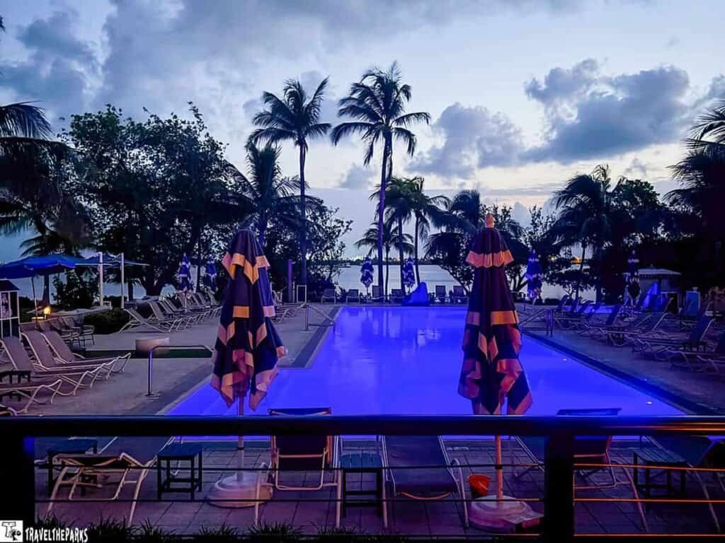 Capitana Resort Key West: A swimming pool at dusk surrounded by palm trees, with striped umbrellas and empty lounge chairs, against a colorful sky.