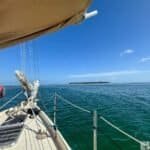 A view from the deck of a Danger Charters sailboat, showing calm turquoise waters and a small green island in the distance under a bright blue sky. Part of the boat's mast and rigging are visible in the foreground.