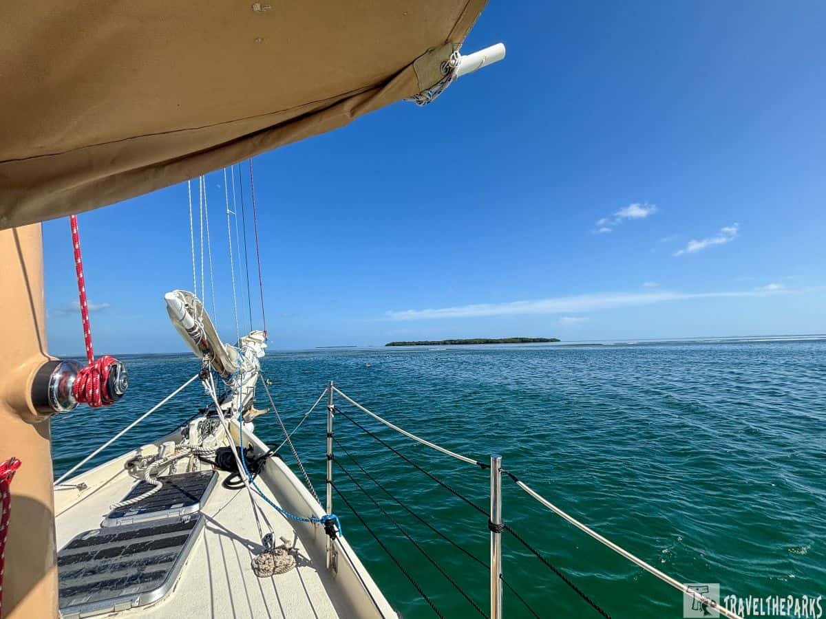 A view from the deck of a Danger Charters sailboat, showing calm turquoise waters and a small green island in the distance under a bright blue sky. Part of the boat's mast and rigging are visible in the foreground.