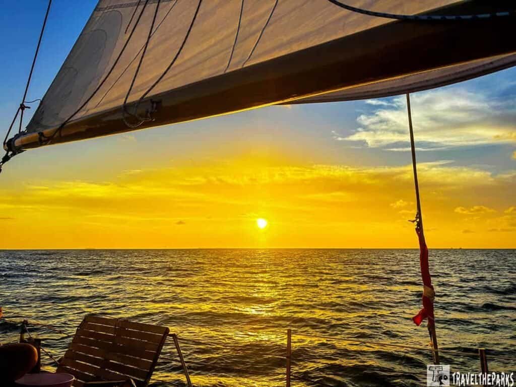 A sunset view from a sailing boat, featuring a vibrant orange and yellow sky, calm ocean waves, and a wooden deck chair.
