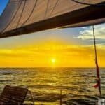 A sunset view from a sailing boat, featuring a vibrant orange and yellow sky, calm ocean waves, and a wooden deck chair.