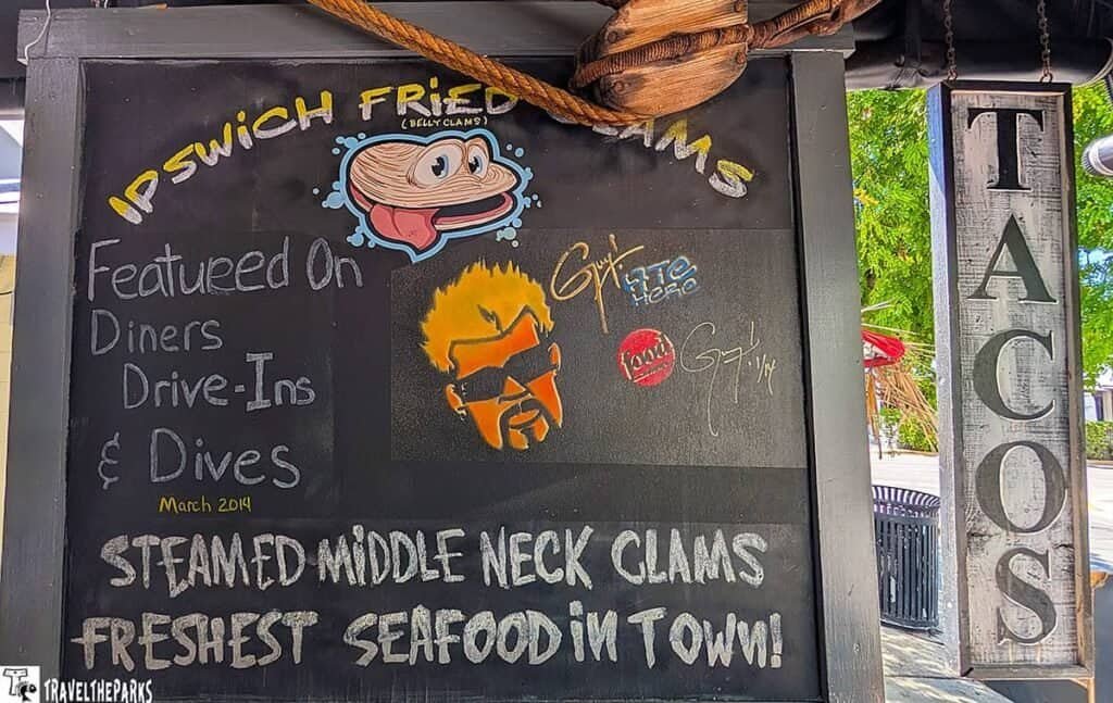 Blackboard menu for "Ipswich Fried Clams" with a cartoon clam illustration, featured on "Diners, Drive-Ins & Dives." Text highlights steamed middle neck clams and fresh seafood. Part of a taco sign is also visible.
Transcribed Text:
IPS WICH FRIED CLAMS
Featured On
Diners Drive-Ins & Dives
March 2014
STEAMED MIDDLE NECK CLAMS
FRESHEST SEAFOOD IN TOWN!