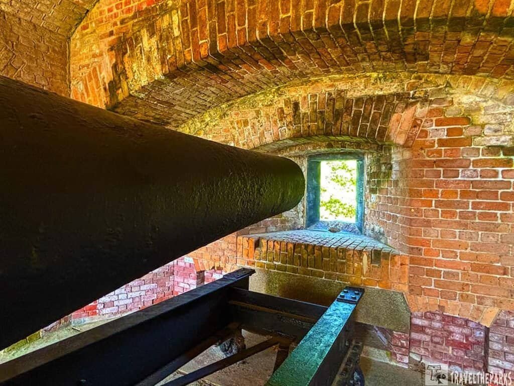 Interior of a historic fort featuring a black cannon pointed towards a stone window framed by red brick walls.