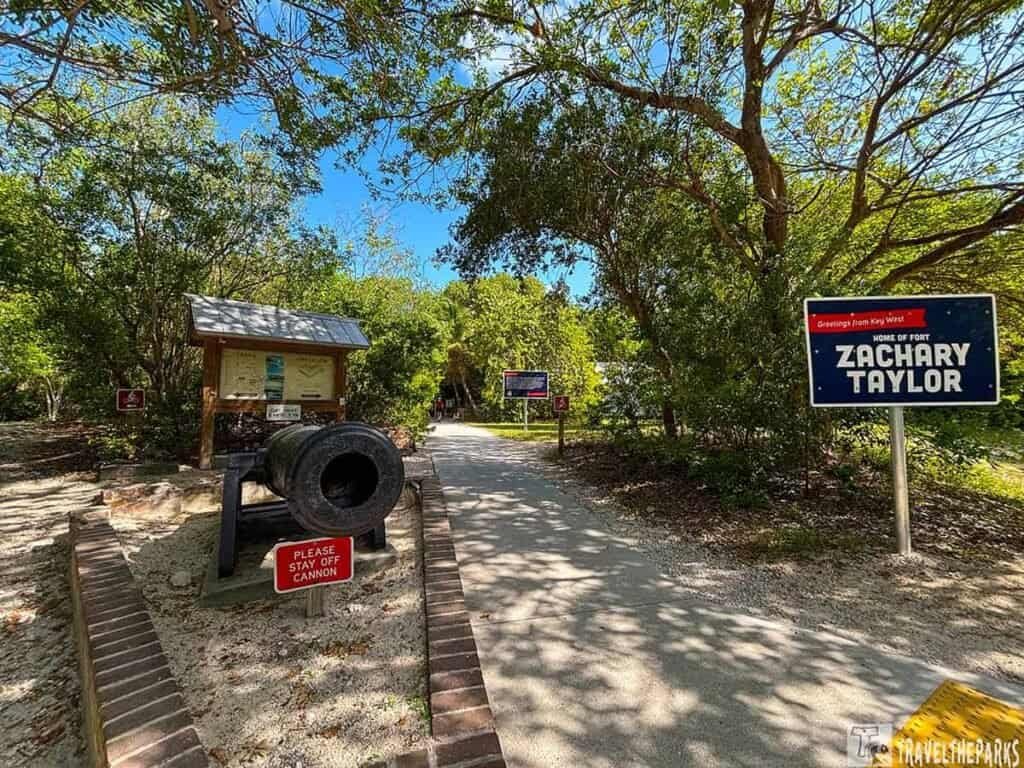 Entrance to Fort Zachary Taylor Historic State Park with a cannon, informational sign, and path lined by trees.