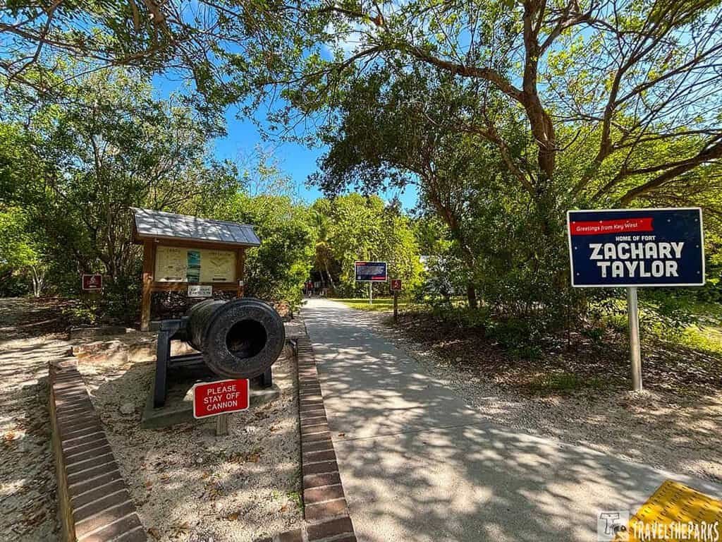 Entrance to Fort Zachary Taylor Historic State Park with a cannon, informational sign, and path lined by trees.