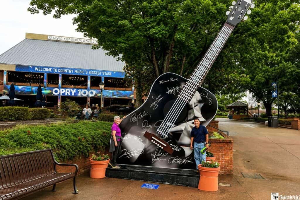 A large black guitar sculpture with signatures stands outside the Grand Ole Opry, with a woman in a pink shirt and a man in a blue shirt posing beside it.

