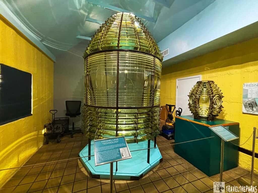 An interior view showing a large Fresnel lens in the center of Key West Lighthouse and Keeper’s Quarters, surrounded by colorful walls and a smaller model to the right.