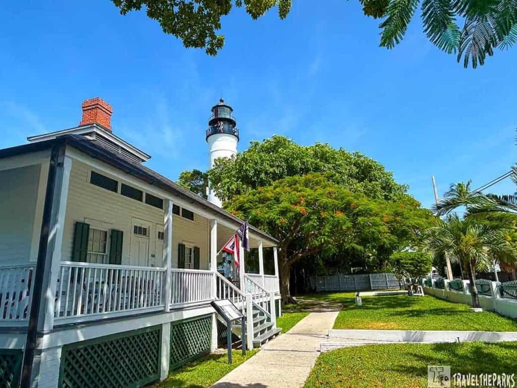 Key West Lighthouse in the background with the white Keeper’s Quarters in the foreground, surrounded by green trees and grass under a blue sky.