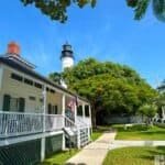 Key West Lighthouse in the background with the white Keeper’s Quarters in the foreground, surrounded by green trees and grass under a blue sky.