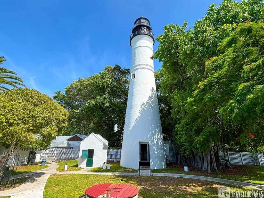 Key West Lighhouse white lighthouse with a black lantern stands amid green trees and a circular red table in a sunny setting.