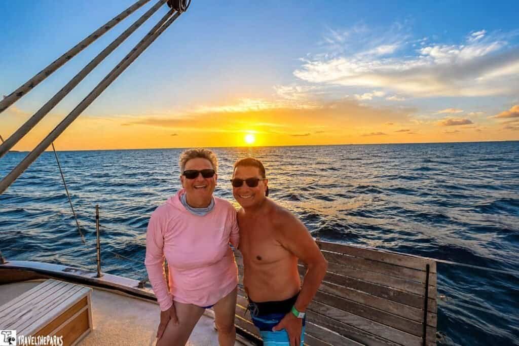 A couple smiles on a DAnger Charters schooner boat during sunset, with a colorful sky and ocean behind them.