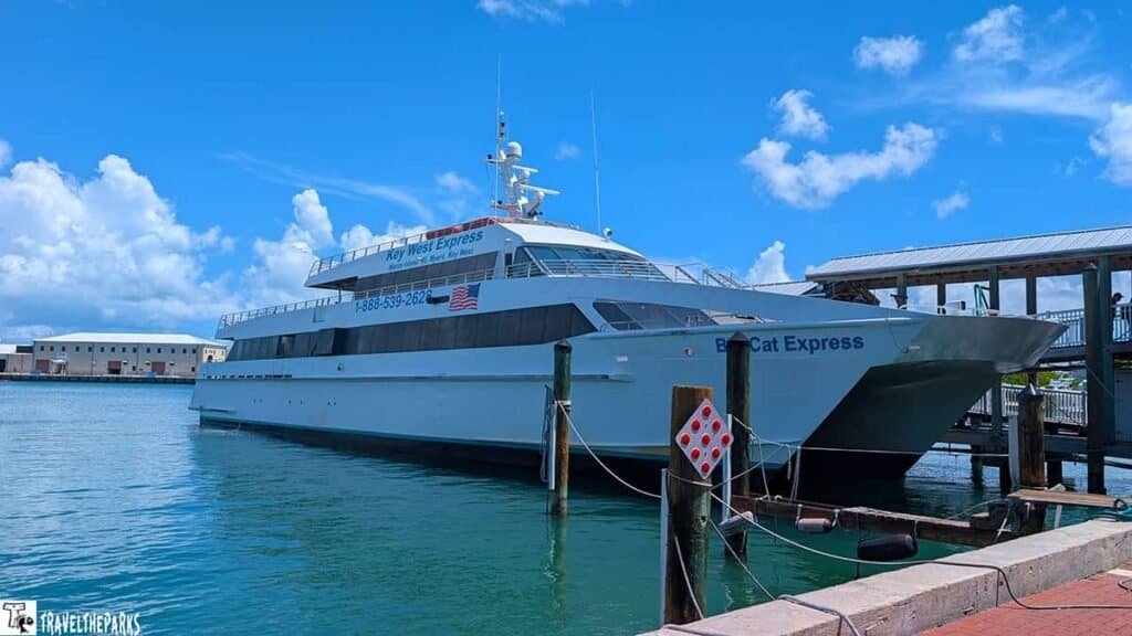 A large white ferry named "Key West Express" is docked in a blue harbor, secured to wooden posts with a sunny sky and white clouds behind it.