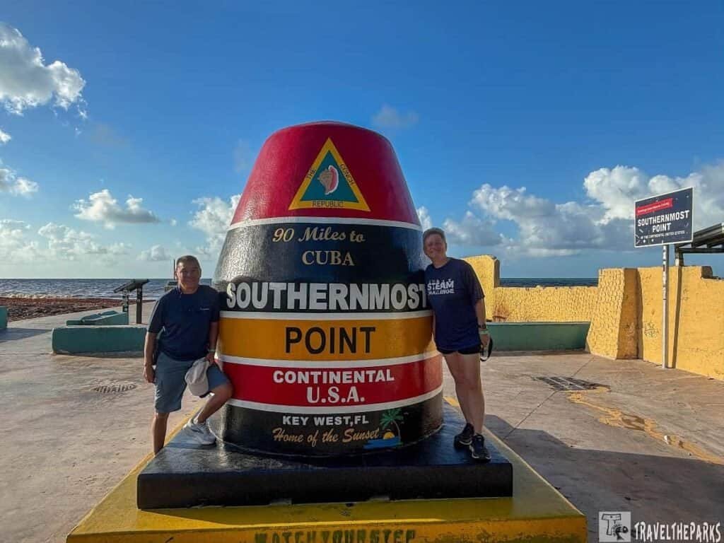 Two people pose beside a colorful buoy marking the Southernmost Point in Key West, Florida, with a blue sky and ocean in the background.