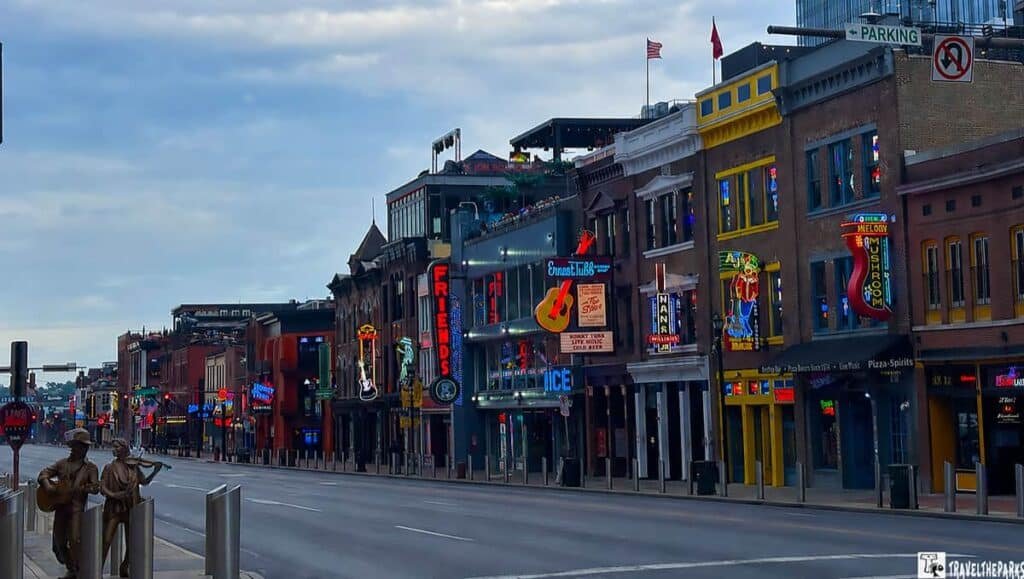 A downtown Nashville Broadway lined with colorful bars and music venues, featuring bronze musician statues in the foreground and a cloudy sky above.