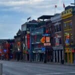 A downtown Nashville Broadway lined with colorful bars and music venues, featuring bronze musician statues in the foreground and a cloudy sky above.