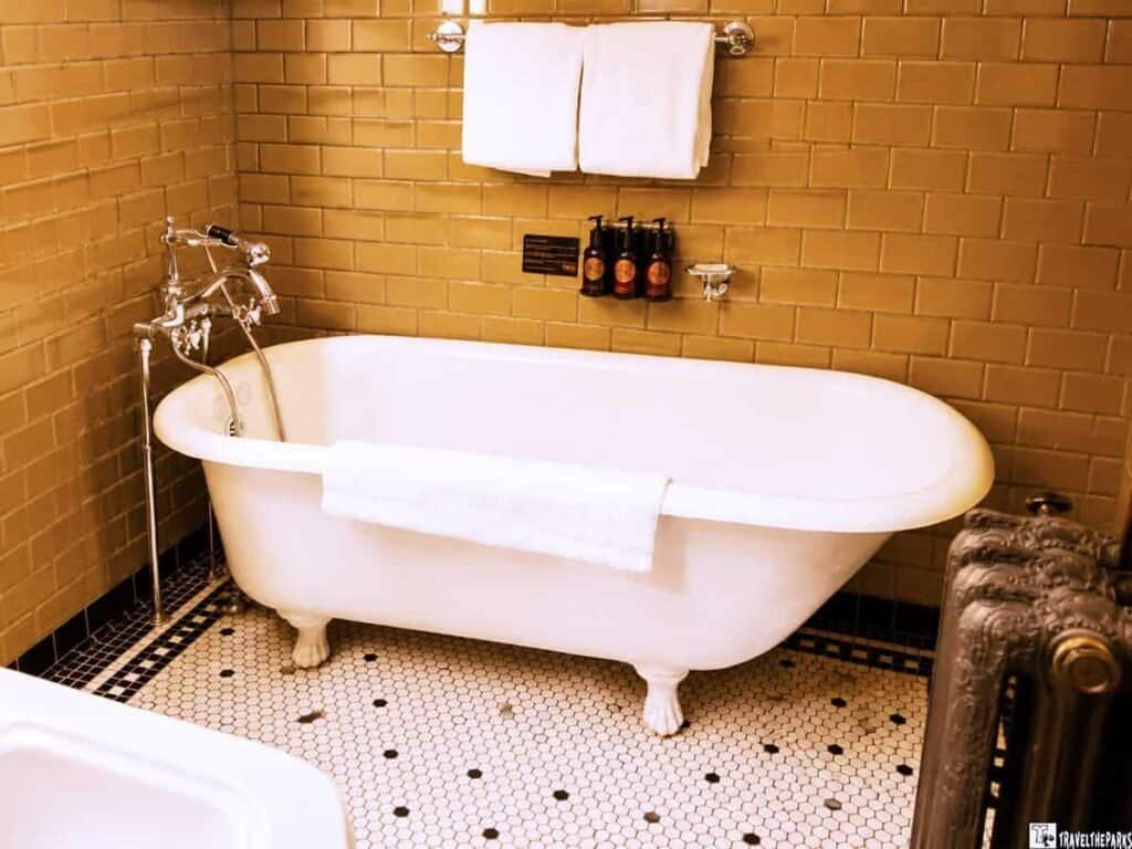 A vintage bathroom featuring a white freestanding bathtub with claw feet, chrome fixtures, and beige subway tile walls. Two white towels hang above the tub, and three dark bottles sit on the wall.