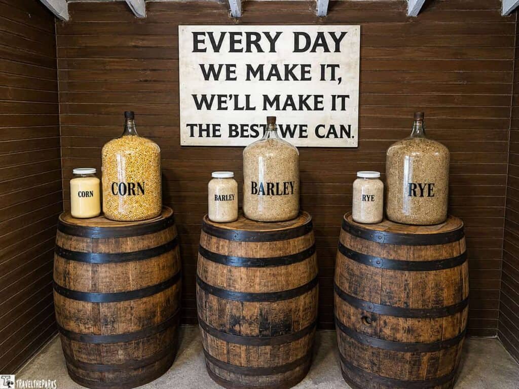Display of three barrels with jars of corn, barley, and rye, accompanied by a sign that reads, "EVERY DAY WE MAKE IT, WE'LL MAKE IT THE BEST WE CAN."