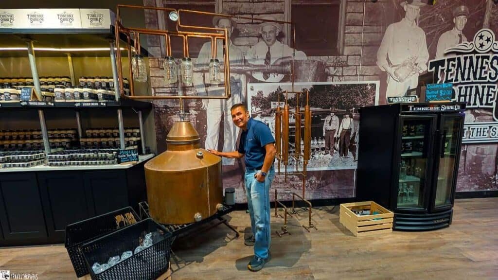 A man stands next to a large copper still in a tasting room, with historical photographs and jars of products in the background.

