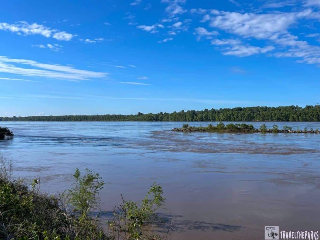 A tranquil Mississippi riverside view at Trail of Tears State Park, featuring a calm brown river bordered by lush green trees under a bright blue sky with scattered clouds.