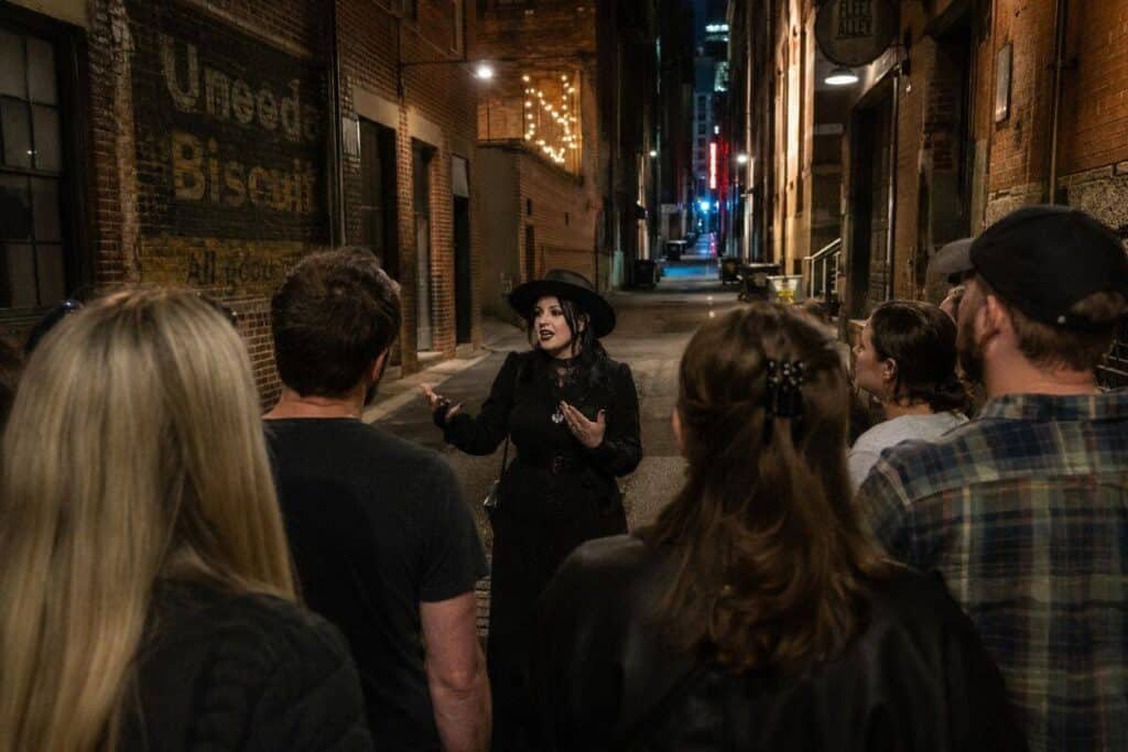 A woman in a black dress and hat speaks to a group in a dimly lit alley, with a faded "Uneeda Biscuit" sign and string lights in the background.

