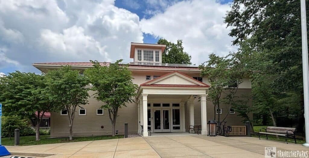 Exterior view of the Ulysses S. Grant Visitor Center, featuring a modern two-story design with a central entrance, white columns, and red roof. Surrounded by green trees and benches.

