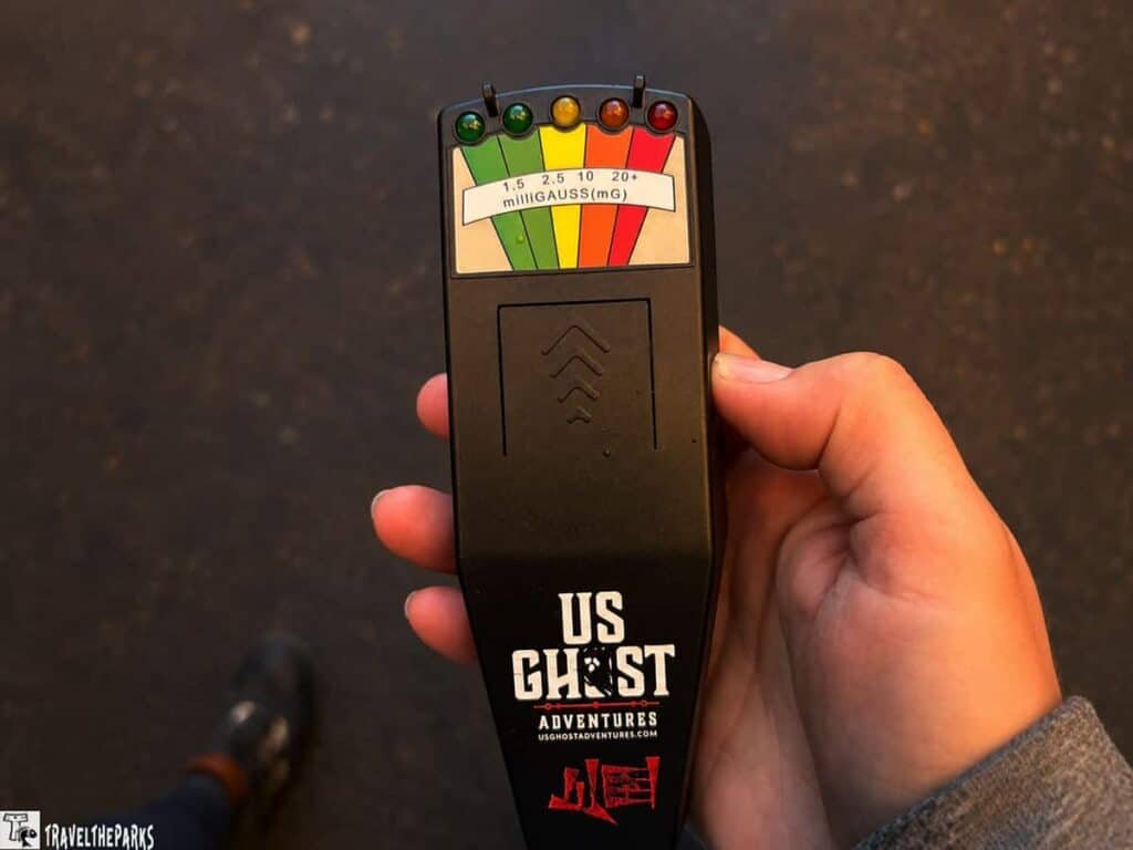 A black handheld ghost detector with a color-coded gauge is held in a person's hand, indicating electromagnetic field levels.

